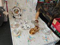Full view of china and glass items on floral sheet including china sets, flower frogs, cut glass jars, Waterford glasses, and wooden pepper grinder