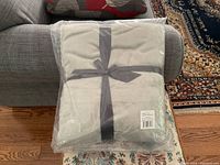 Photo showing the light grey fleece blanket folded and wrapped in clear plastic packaging with a grey ribbon tied around it, placed on a grey fabric couch and patterned carpet.