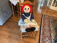Raggedy Ann doll seated on wooden chair, showing full front view, including head, torso, and legs dressed in blue print top, white apron, striped socks, and black shoes.