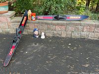 Overall view showing both O'Brien waterskis laid on the ground and propped against a short stone wall. Various boating and decorative items including wooden oar, buoy, and ceramic sailor plaque are positioned nearby.