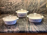 Three Pyrex Corning Ware Cornflower Collection casserole dishes (small, medium, large) with clear glass lids shown on countertop.