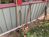 Photo showing five outdoor tools lined up against a building grey wall with red trim: sledgehammer, shovel, axe with broken handle, pitchfork, and another pitchfork or rake