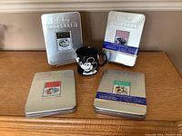 Four silver metal Walt Disney Treasures DVD tins with classic Disney artwork and a black Mickey Mouse ceramic mug on wooden surface
