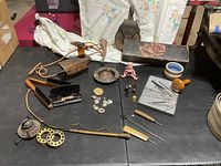 Overview photo showing multiple items on black table including Victorian whip, miniature doll, copper ashtray, antique box, eyeglasses with case, folk art piggy bank, pulley, musical instrument, sewing tools and pins.