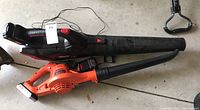 Two cordless leaf blowers on a concrete garage floor. One is black with red details (Walmart TC20BLB) with battery attached. Second is orange (Black & Decker) with 20V Lithium Ion battery attached.