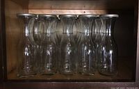 Six clear glass bottles with slightly flared tops standing upright in a wooden shelf.