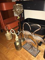Lot showing two brass crane bird figurines, a 20-inch brass candle holder with crystal embellishments, and a brass bell with rope handle on a wooden floor.