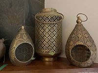 Three brass lanterns displayed side by side: two teardrop-shaped with cutout designs and one cylindrical with geometric pattern and handle.