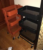 Photo showing both black and rust orange rolling organizer carts side by side on wooden floor, each with three tiers of mesh baskets on wheels.