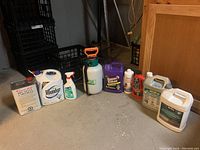 Full view of cleaning supplies including Roundup, Super Clean, a sprayer and other cleaners all placed on basement concrete floor.