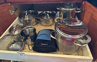 Cookware set arranged in a kitchen drawer showing pots, pans, lids, and accessories including silicone mitts.