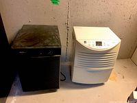 Photo showing side-by-side black Danby and white GE dehumidifiers on basement floor with visible dust.