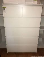Front view of white IKEA style dresser with five drawers, standing between shelves.