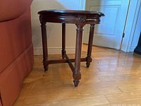 Side view of the wooden end table showing carved legs and stretcher base on hardwood floor near door and couch.
