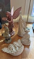 Photo of five resin figures on wooden floor: large pink-winged fairy, white angel, seated Buddha, two cherub heads ornament