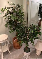 Two faux trees next to each other inside a room, one in a wicker basket pot, the other in a ceramic pot alongside two white lamps.