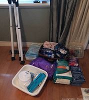 Overview of healthcare supplies and security bars arranged on floor showing various packaged medical and hygiene items, a white tub holding medical components and three white adjustable security bars leaning against a wall.