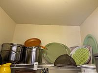 Two large stainless steel pots, a copper-colored frying pan, black wire mesh strainer, green plates or serveware, and table tent on a shelving unit.