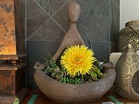 Full view of the concrete Namaste pose planter showing the entire figure with artificial succulents and yellow flower in the basin.