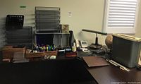 Office desk surface displaying mailbox-style black plastic sorters, pen holders, rotary phone, green lamp, globe, shredder and organizers.