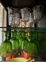 Overall set showing six green pressed glass goblets upside down on the bottom shelf and four clear glass etched stemware and one pitcher on top shelf