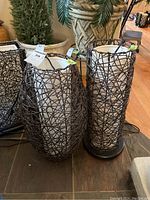 Pair of two woven string lamps of different sizes sitting on floor with plant pots in background