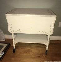 Photo of front view of the white tea trolley cart with folded sides, showing floral carved detailing and painted surface.
