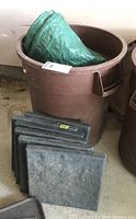 Brown Rubbermaid trash barrel with green tarps inside and six gray resin stone tiles standing beside it on the floor