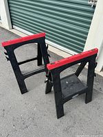 Pair of black plastic collapsible work horses with red tops side by side on pavement outside storage units.