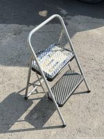Front and side view of a silver metal 2 step ladder with black slip-resistant bottom step and diamond plate top step, standing outdoors on pavement.