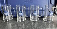 Four clear pint-sized beer mugs with Toronto Maple Leafs logos arranged in a row on a dark surface