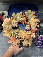 View of autumn wreath showing large maple leaves, berries, twigs, and small pumpkins, held against a blue background with a soda can for scale.