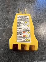 Photo showing the A.W. Sperry Instruments circuit receptacle tester with yellow plastic body, indicator lights, and wiring fault chart label.
