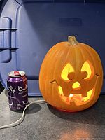 Front view of the electric pumpkin lit up with light shining through carved facial features beside a soda can for scale.