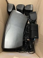 Photo of six speakers and speaker wires in a cardboard box, showing one large JBL center channel speaker and five smaller Bose satellite speakers with wires.