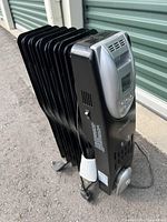 Black Noma electric oil-filled radiator heater with digital control and seven fins, standing on an outdoor surface against a green door background.