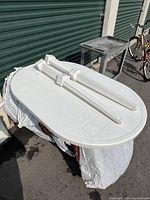Photo of the white oval plastic resin patio table top with four detachable legs positioned on top, displayed outdoors on a makeshift support.