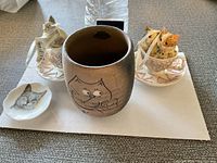 Two cat figurines on saucers, ceramic cup with cat face design, and small cat dish.