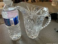 Clear crystal pitcher beside a water bottle for size comparison. Pitcher features detailed cut glass pattern and a handle.