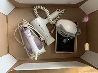 Eye massager, Braun hair dryer, and steam iron inside a box, showing coiled cords and accessories.