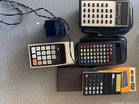 Top-down photo of four vintage calculators together on carpeted surface showing the various colors, buttons, and sizes including the DGT 1000 in black with orange and white keys.