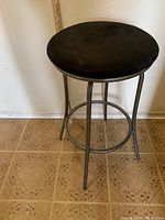 Photo of a black high stool with a soft round seat and metal frame against a plain wall.