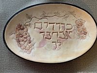 Full view of the oval ceramic Judaica plate with Hebrew inscription in the center and grapevine clusters along the sides. Beige color with raised design and dark rim.