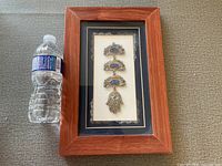 Full view of framed Judaica piece with Hamsa hand and Hebrew script plaques, alongside a water bottle for size reference.