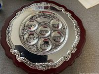 Top front view of the Passover Seder plate showing the silver-tone metal plate with Hebrew labeled sections on a wooden base with scalloped edges.