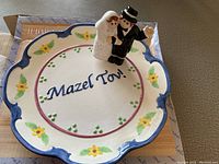 Front view of the wedding plate with 'Mazel Tov!' inscription and floral border, with bride and groom figurine placed on the plate.