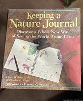 Front cover of the book 'Keeping a Nature Journal' showing artistic nature journal sketches and supporting accessories.