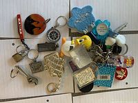 Top view photo of assorted keychains laid out on a white tiled surface showing diverse figures and shapes including animal, promotional items, and miniature souvenirs.
