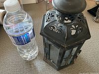 Side view of black metal lantern next to a water bottle for size comparison, showing oxidation and glass panels.