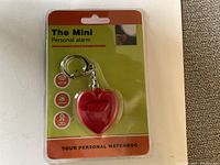 Front of package showing red heart-shaped mini personal alarm with keyring, sealed in plastic packaging.